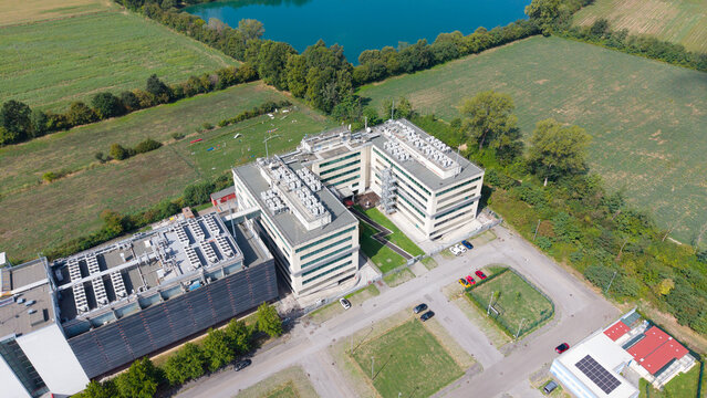 Aerial view of a modern data center complex with rooftop cooling units, surrounded by green fields and a small lake in Settimo Milanese, Lombardy, Italy.
