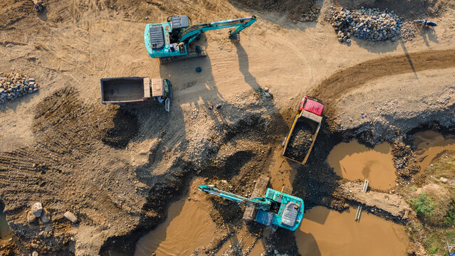 Aerial view of construction site with two excavators and dump trucks working on muddy ground with pools of water and piles of rocks in Karanglewas, Central Java, Indonesia.