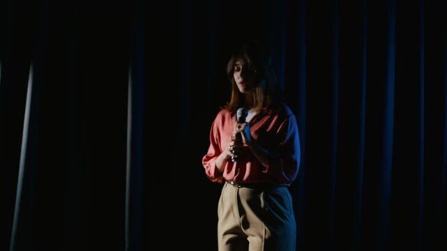 Caucasian young woman stepping onto theater stage into spotlight, holding microphone and addressing audience. Footage suits public speaking training, drama school promotions and arts projects