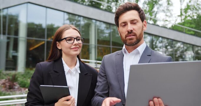 Portrait of businessman and businesswoman standing outdoors, looking at a laptop screen and having a business conversation.