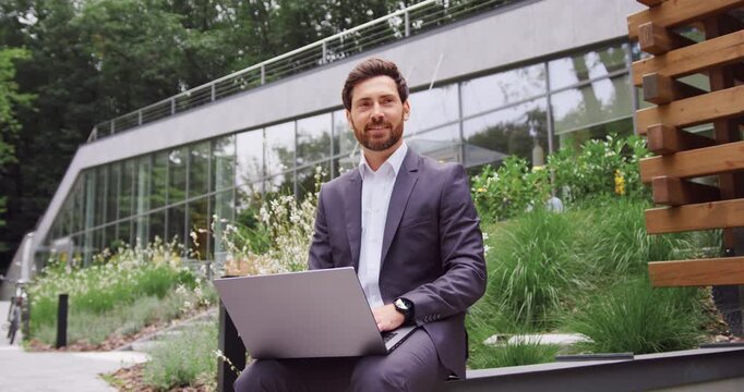 Handsome bearded businessman in a suit sitting on a bench in a city park, working on a laptop and thoughtfully looking into away.