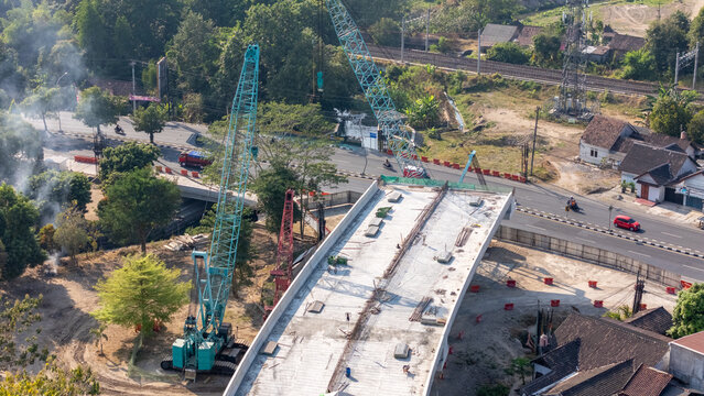 Aerial view of a bridge construction site with large cranes, concrete structures, and surrounding roads and houses in Yogyakarta, Special Region of Yogyakarta, Indonesia.