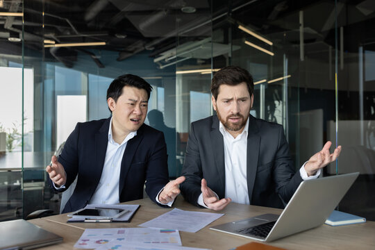 Two businessmen at an office desk react with frustration and worry as they review a laptop showing bad financial charts, reports and documents during a tense meeting