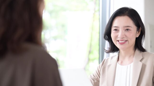 A middle-aged woman smiling as she meets with a client for a face-to-face consultation by a window surrounded by fresh greenery; a businesswoman giving explanations and making proposals in an office&mdash;v