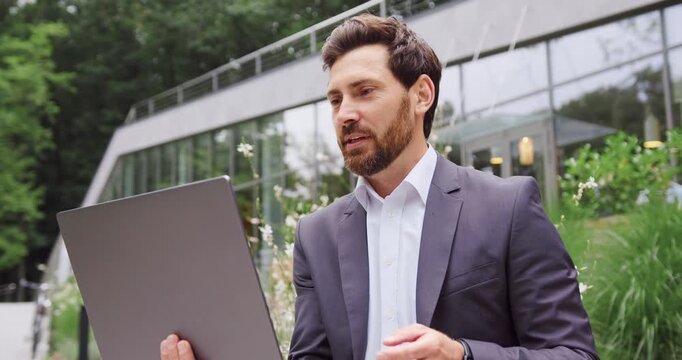 Businessman sitting outdoors in a city park, working on a laptop and having a professional video call while looking at the screen and greeting a business partner.