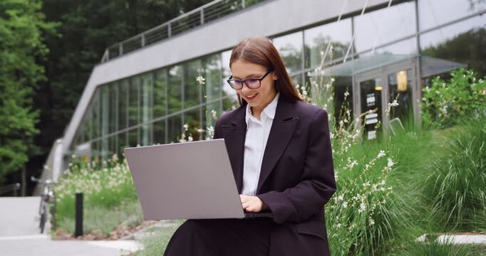 Young businesswoman sitting on a bench in a city park, typing on a laptop and working outdoors while focused on the screen.