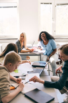 Happy teenage girl talking with friends while sitting at desk in high school classroom