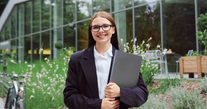 Portrait of a young businesswoman in a professional business suit standing outdoors with a laptop, looking at the camera and smiling against the background of a modern glass office building.