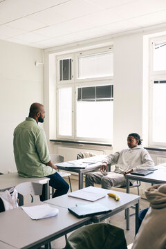 Male teacher talking with teenage boy sitting at desk in high school classroom