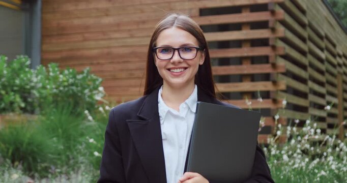 Young woman in glasses posing outdoors in a city park, looking to the side and then smiling at the camera.
