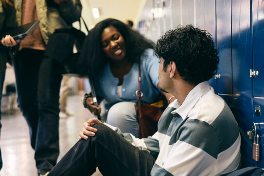Smiling teenage girl consoling male friend sitting alone near lockers in school corridor