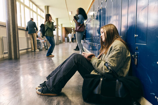 Teenage girl using smart phone while sitting on floor near lockers in school corridor