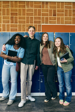Full length portrait of smiling male and female teenage students standing with arm around each other near locker in scho