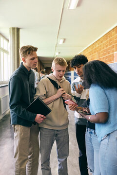 Teenage girl showing smart phone to friends while spending leisure time in college corridor