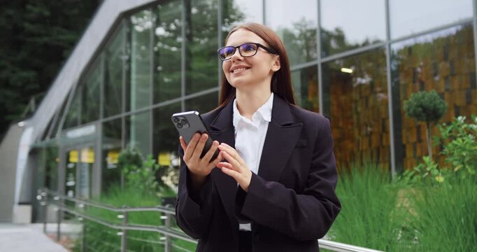Young woman in formal business attire holding a mobile phone and looking to the side against the background of a modern glass office building.
