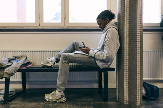 Full length of teenage boy using smart phone while sitting on bench at school corridor