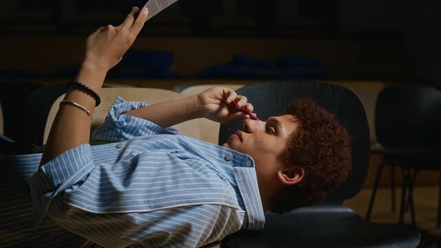 African American teenage actor lying on chairs reading script and rehearsing lines in student theater. Content perfect for drama education campaigns and youth arts promotion