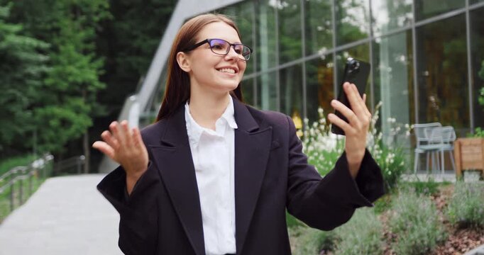 Young woman greeting a colleague with a friendly wave while walking through a city park and talking on her smartphone.