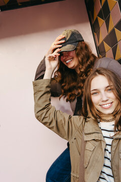 Portrait of smiling woman holding cap of her female friend standing near wall