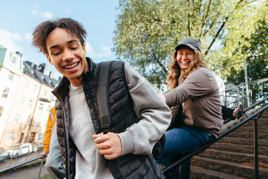 Happy teenage boy holding backpack strap while having fun with female friend sitting staircase railing