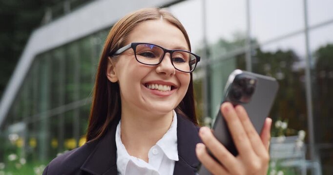 Close-up portrait of a young woman smiling with joy as she looks at her smartphone screen, reacting happily to something she sees on her phone.