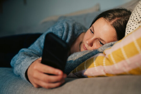 Teenage girl scrolling social media on her smart phone while lying down on couch