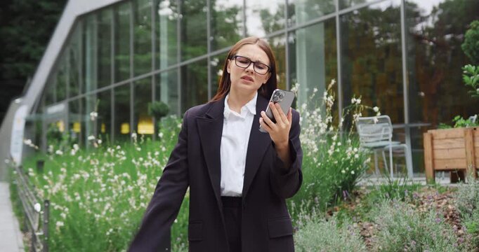 Young Caucasian woman walking in a city park, looking disappointed and concerned as she reads an unexpected message on her smartphone.