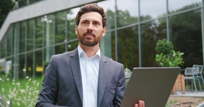 Portrait of businessman sitting on a bench in a city park, working on a laptop outdoors, focused on the screen before briefly looking away to the side.
