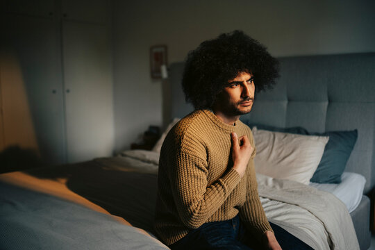 Thoughtful man with curly hair sitting on bed at home