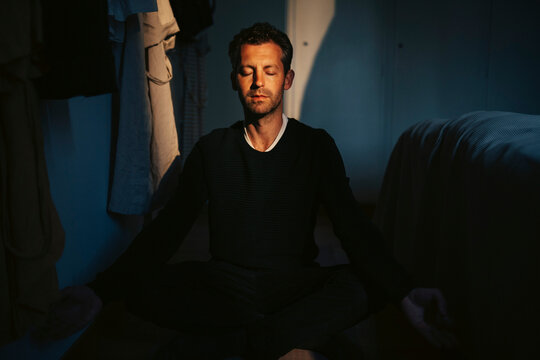 Man with eyes closed meditating while sitting on floor in dark room