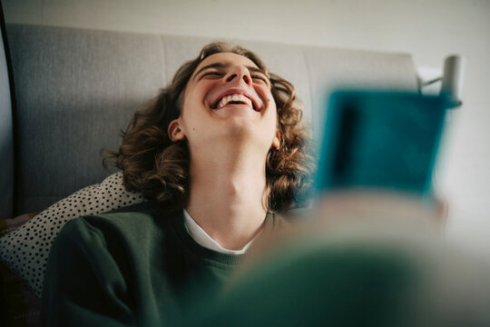 Cheerful man holding phone and laughing while sitting on bed