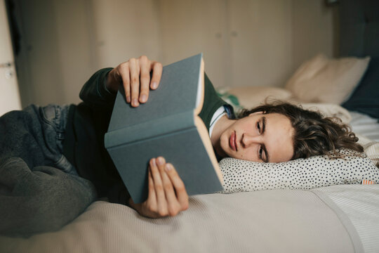 Young man spending leisure time reading book while lying on bed at home
