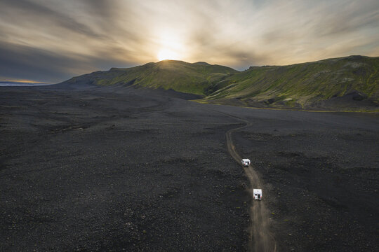 Aerial view of two white camper vans driving along a winding dirt road through a vast volcanic landscape with green hills under a dramatic sunset sky in Iceland.
