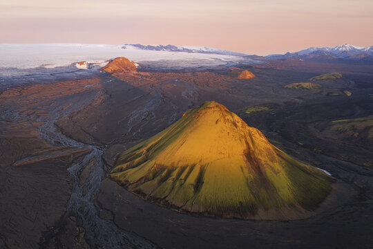 Aerial view of Maelifell volcano with its vibrant moss-covered slopes set against a vast volcanic landscape and distant glacier under soft golden hour light in Skaftarhreppur, Iceland.