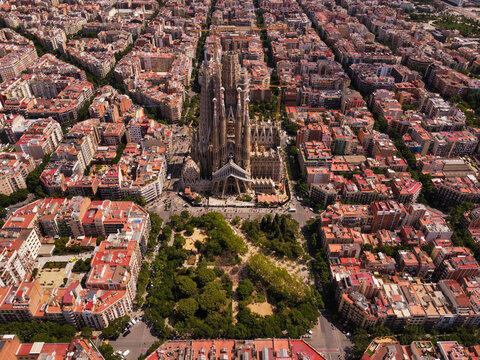 Aerial view of Sagrada Familia surrounded by the dense urban grid of the Eixample district with lush park greenery and historic architecture in Barcelona, Catalonia, Spain.