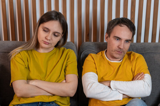 Young married couple sitting on a sofa with crossed arms, refusing to talk to each other. Family conflict, resentment, and silent treatment.