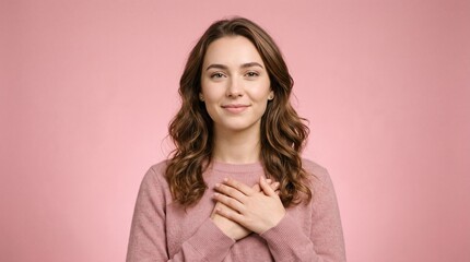 Fototapeta premium Portrait of a thankful smiling young woman holding hands on her chest against pink background