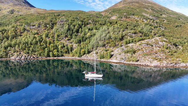 Aerial view of a sailboat floating on the calm blue water of a fjord surrounded by green hills and rocky mountains Skjervoy, Troms, Norway.