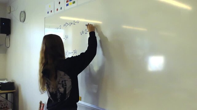 Young female student with long hair writing a complex mathematical formula on a whiteboard in a classroom. Educational concept of a smart pupil thinking and solving an equation during a lesson