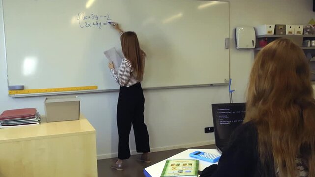 Female mathematics teacher writing a system of linear equations on a whiteboard while a student uses an artificial intelligence program on a laptop to find the solution