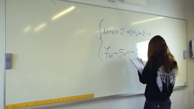 Young female student with long hair writing a mathematical formula on a large whiteboard in a classroom while holding a notebook, demonstrating problem solving and educational concepts