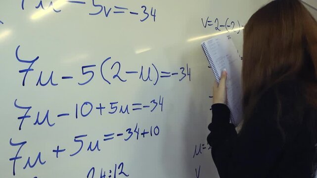 Young female student holding a notebook and writing a mathematical equation on a whiteboard with a marker, solving a complex algebra problem during a lesson in a school classroom