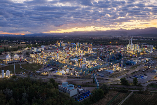 Aerial view of the illuminated industrial plant in Bantzenheim with tall chimneys, storage tanks, and complex infrastructure under a dramatic sunset sky in Bantzenheim, Grand Est, France.