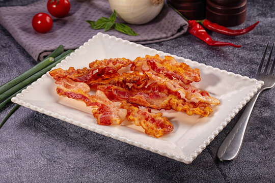 Crispy fried bacon strips on white rectangular plate with fresh vegetables and fork on dark stone background