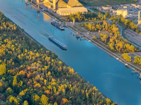 Aerial view of cargo barges navigating the Grand Canal d'Alsace near industrial facilities surrounded by lush autumn forests in Bantzenheim, Grand Est, France.