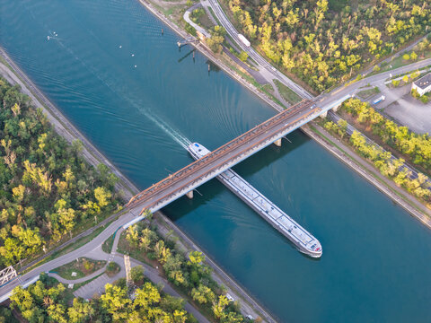 Aerial view of a cargo ship navigating a canal under a bridge surrounded by lush green trees and vegetation in Bantzenheim, Grand Est, France.