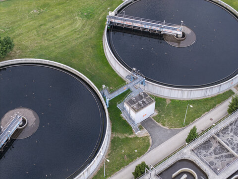 Aerial view of a sewage treatment plant with large circular sedimentation tanks and industrial infrastructure surrounded by green grass in Poland.