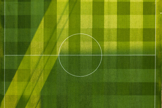 Aerial view of a soccer field with a distinct checkered grass pattern and white markings, illuminated by sunlight casting long shadows across the pitch in the United Kingdom.