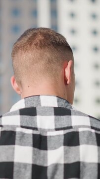 Close up of man with short hair wearing black and white checkered shirt seen from behind. background shows urban setting with tall buildings visible through soft daylight lighting. scene