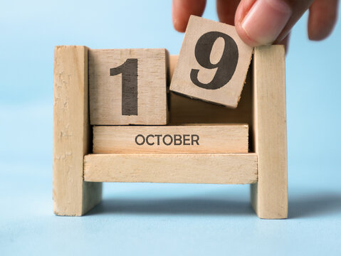 A woman's hand is holding a wooden block with the number 9 written on it on a wooden block calendar for October 19th on a light blue background.
